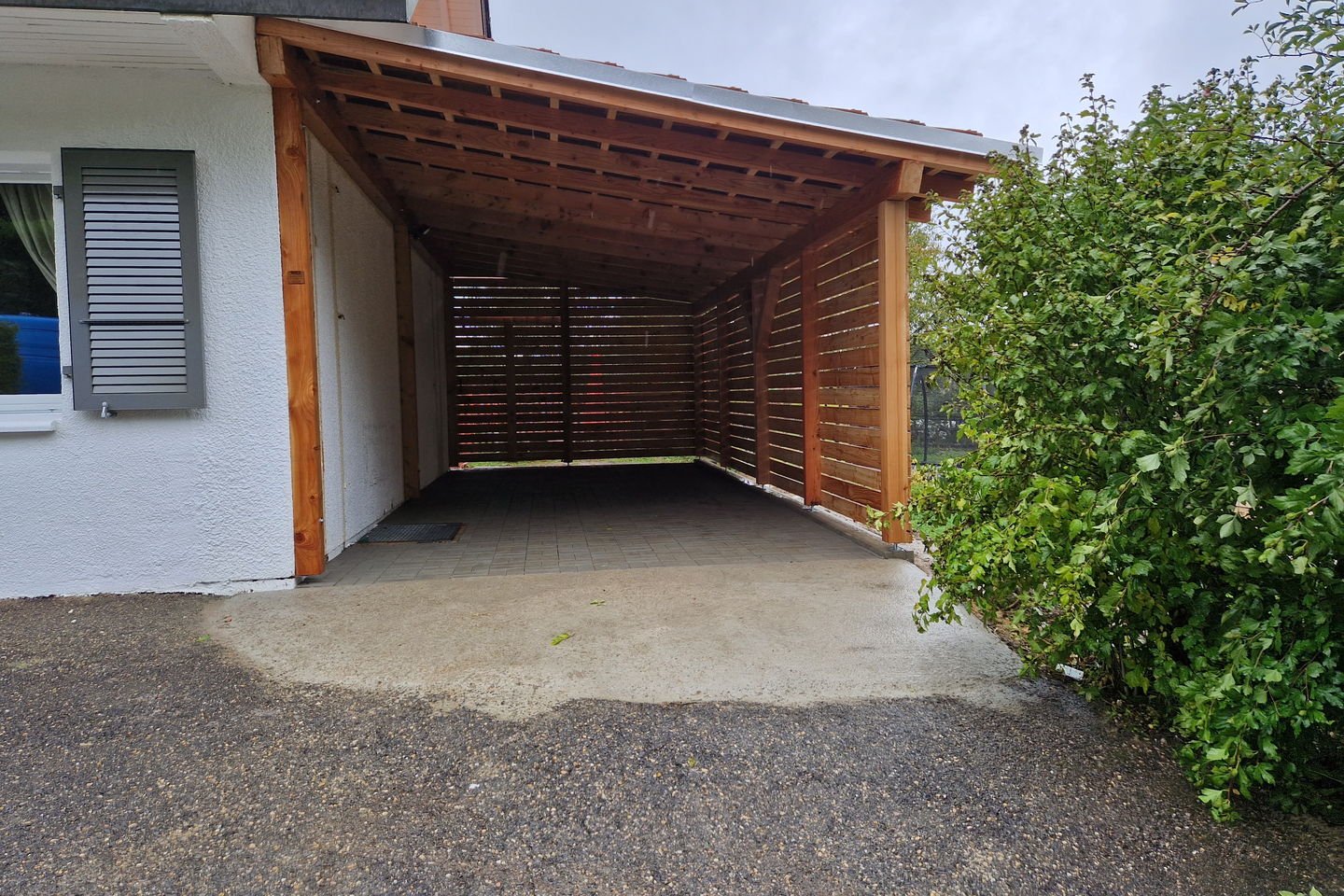 Couvert à voitures en bois adossé à une maison blanche, avec sol en gravier stabilisé
