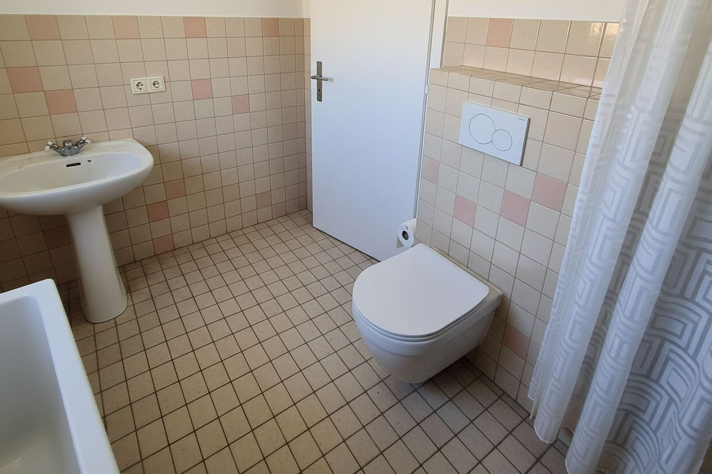 Salle de bains reconstituée avant travaux : petits carreaux beiges et roses, lavabo sur colonne, rideau de douche