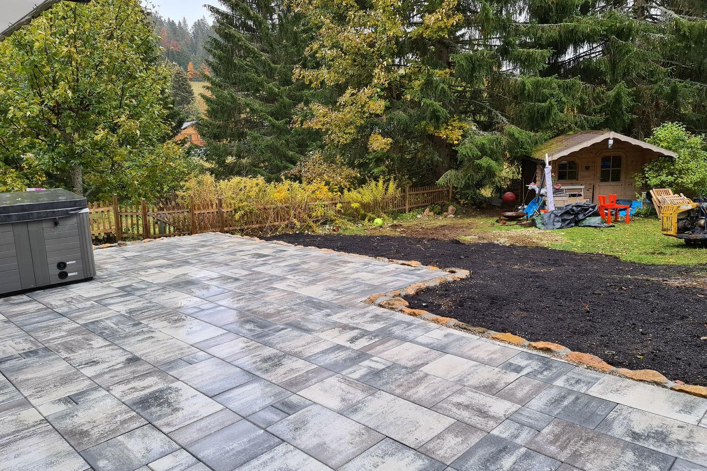 Vue large de la terrasse en dalles avec le jardin, les sapins et un cabanon en arrière-plan