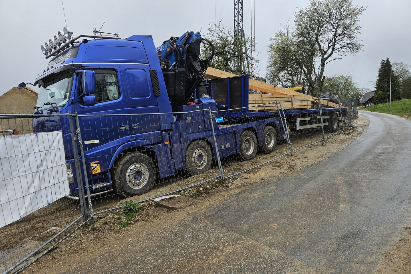 Camion-grue bleu chargé d’une charpente en bois, arrêté sur une route de campagne étroite